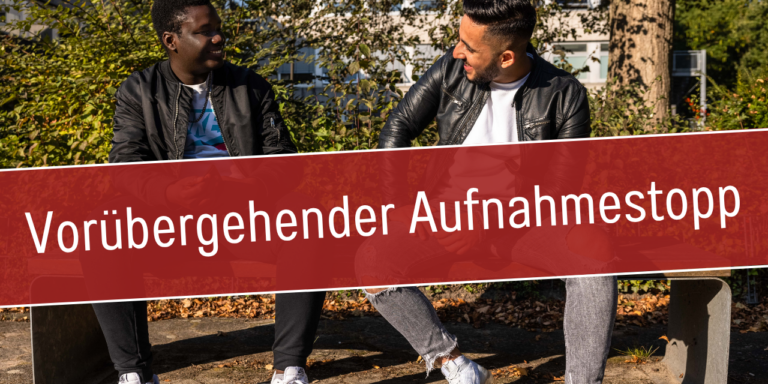Zwei junge Männer sitzen lachend auf einer Bank im Freien; Schriftzug: Vorübergehender Aufnahmestopp