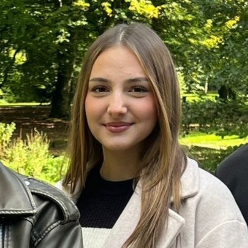 Junge Frau mit langen braunen Haaren lächelt vor grünem Parkbäumen im Hintergrund