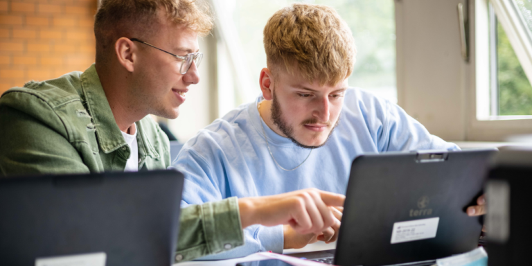 Zwei junge Männer arbeiten gemeinsam an einem Laptop in einem hellen Klassenraum