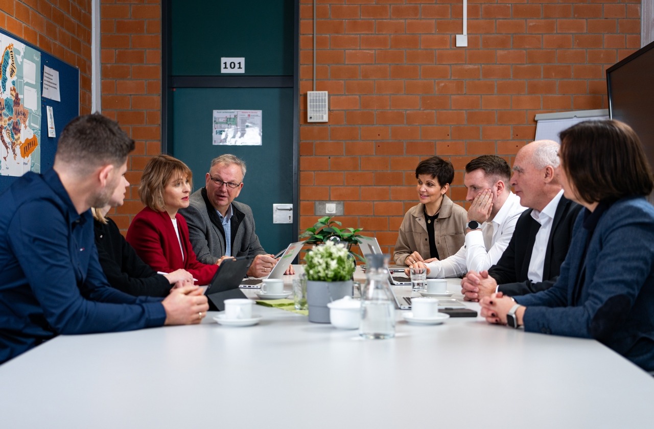 Gruppe von sieben Personen in einer Besprechung an einem weißen Konferenztisch in einem Raum mit Backsteinwand