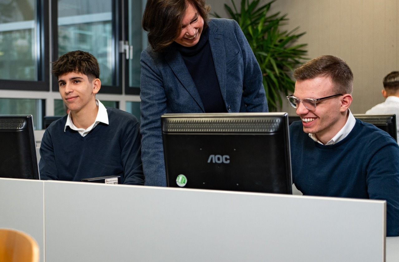 Eine Lehrerin beugt sich lächelnd über zwei junge Männer, die an Computerarbeitsplätzen sitzen und lachen