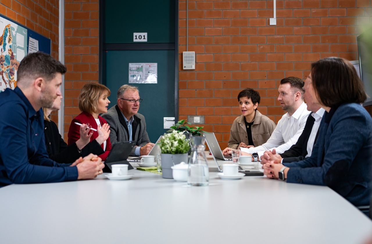 Gruppe von sieben Personen in einer Besprechung an einem Konferenztisch mit Laptops in einem Büroraum mit Backsteinwand