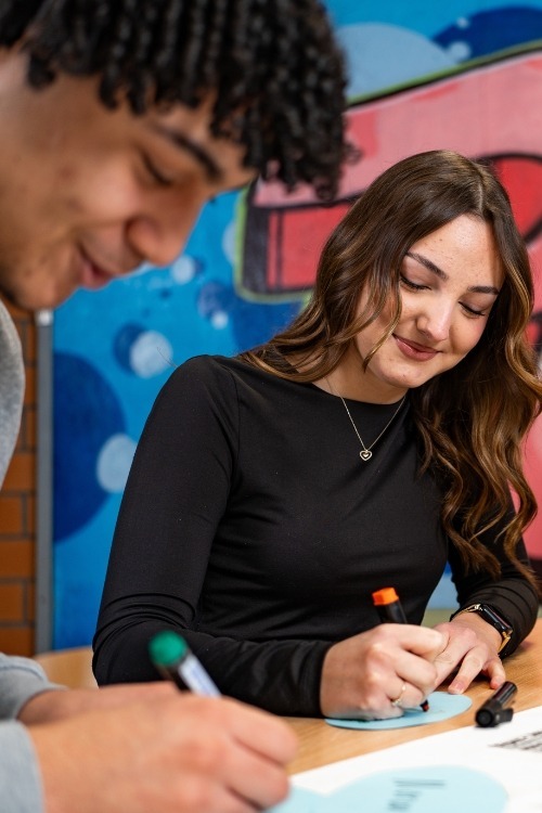 Zwei junge Menschen arbeiten gemeinsam an einem Tisch mit Stiften, im Hintergrund ein buntes Graffiti