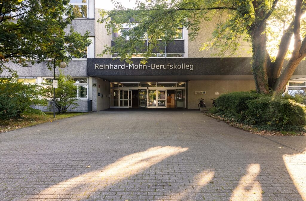 Eingangsbereich des Reinhard-Mohn-Berufskollegs mit gepflastertem Vorplatz und Bäumen