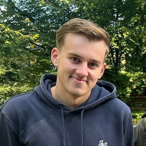 Junger Mann mit kurzem Haar und dunklem Hoodie lächelt vor grünem Baumhintergrund