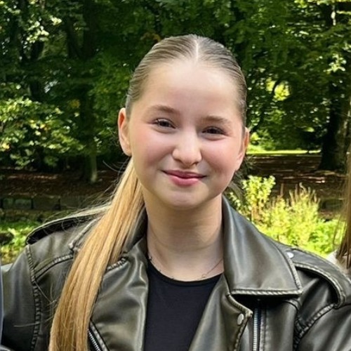 Junges Mädchen mit langen blonden Haaren und schwarzer Lederjacke lächelt vor grünem Baumhintergrund