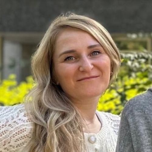 Porträt einer lächelnden Frau mit langen blonden Haaren vor einem unscharfen Hintergrund mit Grünpflanzen