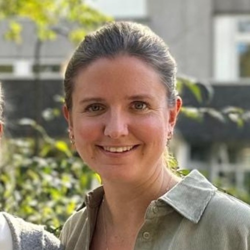 Lächelnde Frau im Freien vor grüner Vegetation und einem Gebäude im Hintergrund