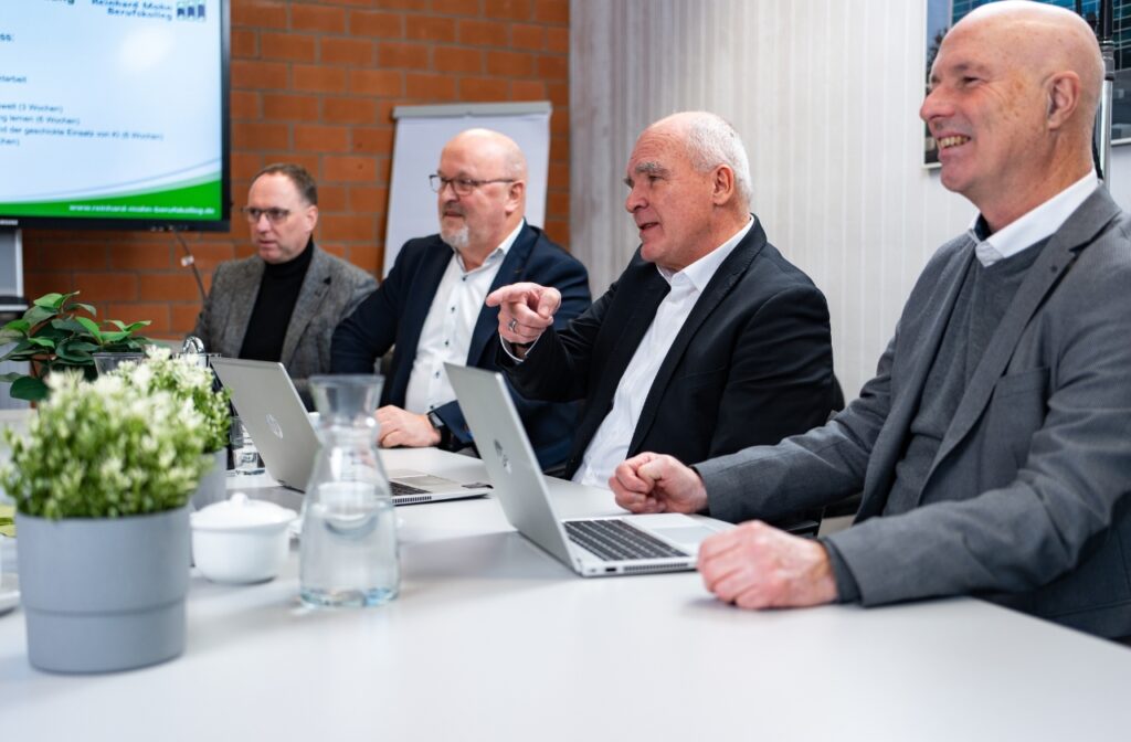 Vier Männer sitzen an einem Konferenztisch mit Laptops; einer spricht und zeigt mit dem Finger, im Hintergrund eine Präsentation des Reinhard-Mohn-Berufskollegs