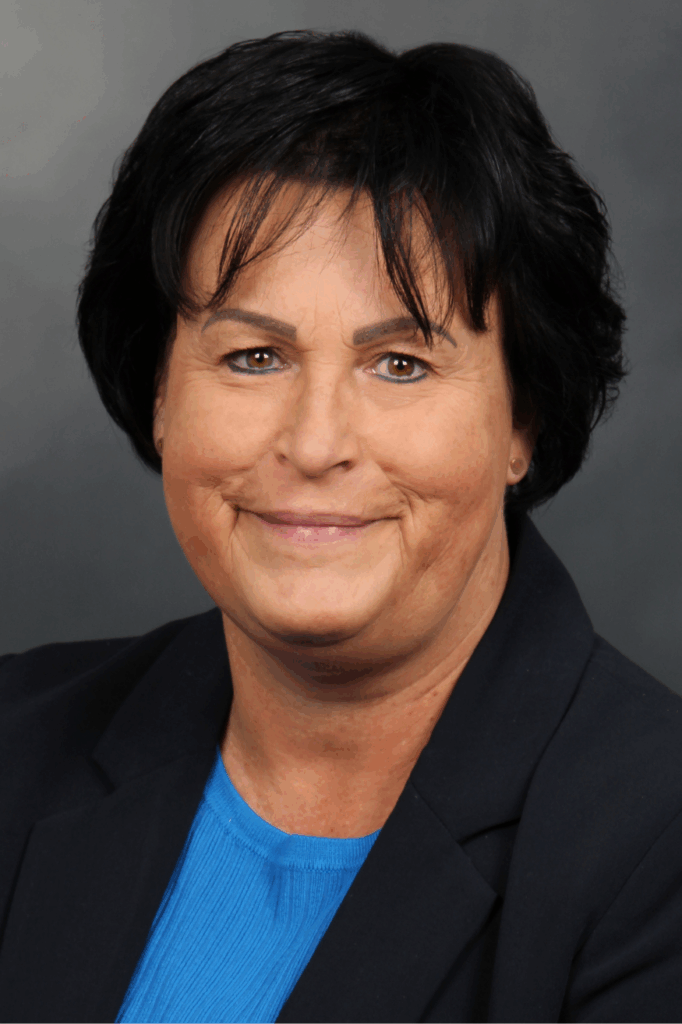 Professionelles Porträt einer lächelnden Frau mit schwarzem Haar, schwarzem Blazer und blauem Oberteil