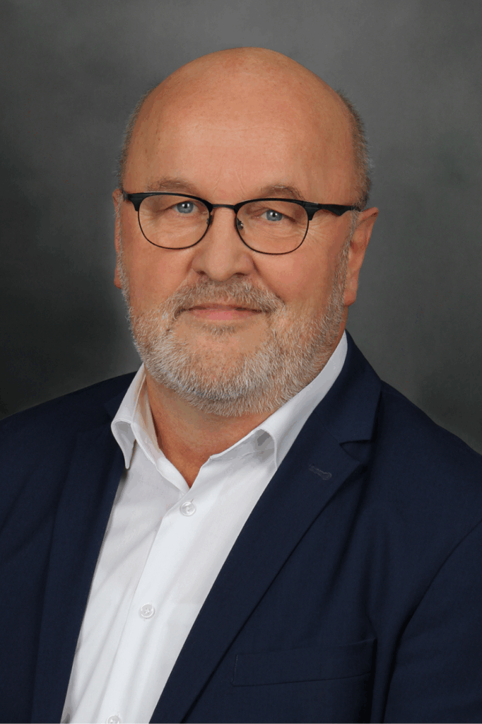 Porträt eines älteren Mannes mit Brille, grauem Bart und blauem Sakko vor grauem Hintergrund