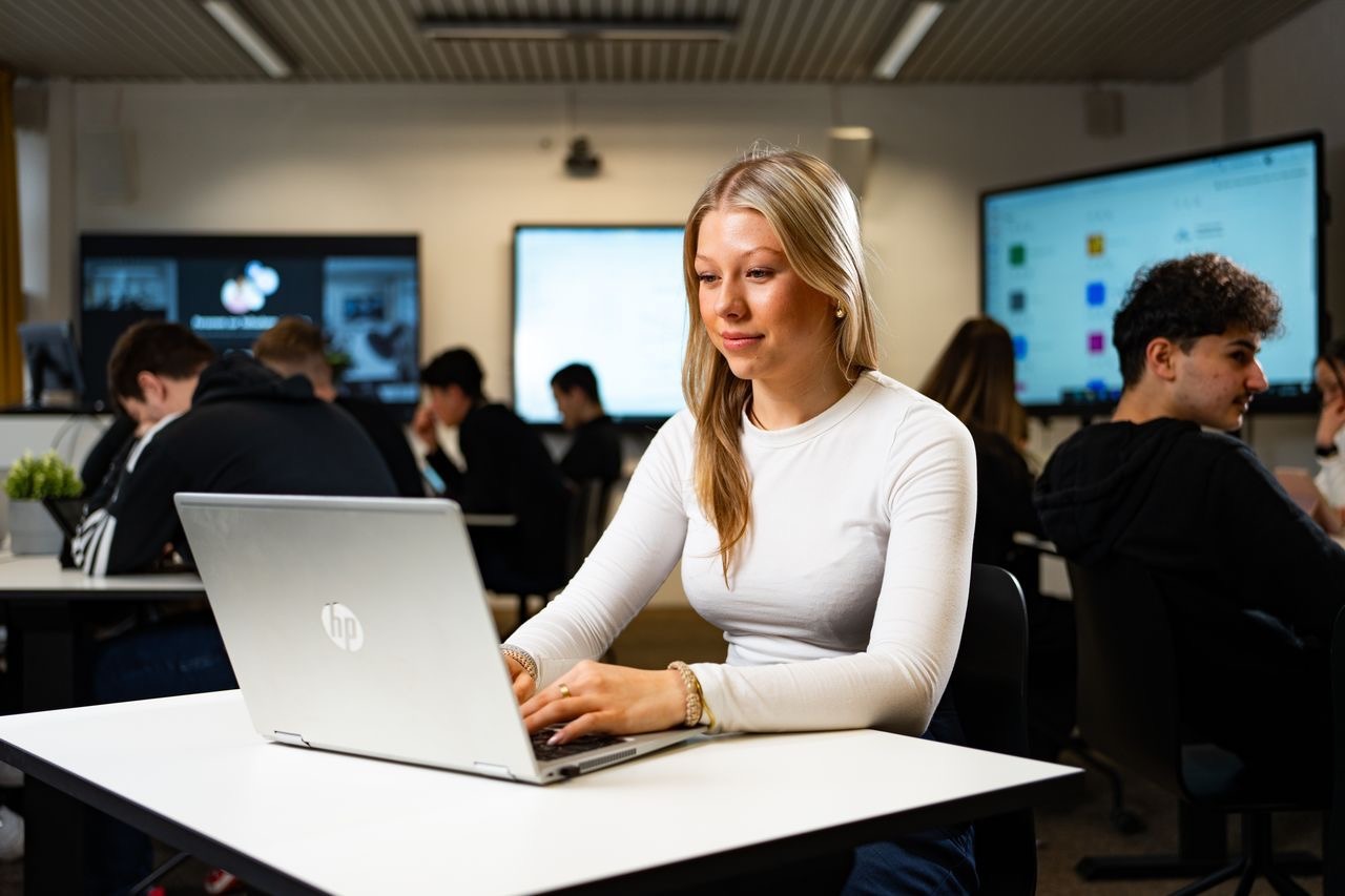 Studentin arbeitet an einem HP-Laptop in einem modernen Klassenraum mit weiteren Studierenden im Hintergrund