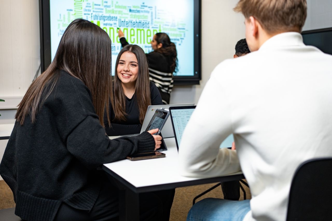Studierende arbeiten mit Laptops und Tablets in einem Seminarraum; im Hintergrund zeigt ein Bildschirm eine Wortwolke zum Thema Management