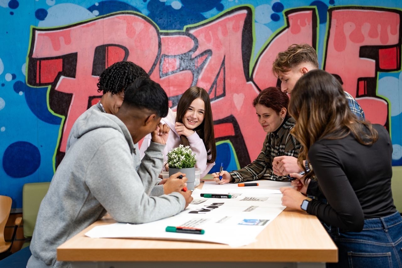 Jugendliche arbeiten gemeinsam an einem Tisch mit Markern und Papieren, im Hintergrund ein buntes Peace-Graffiti