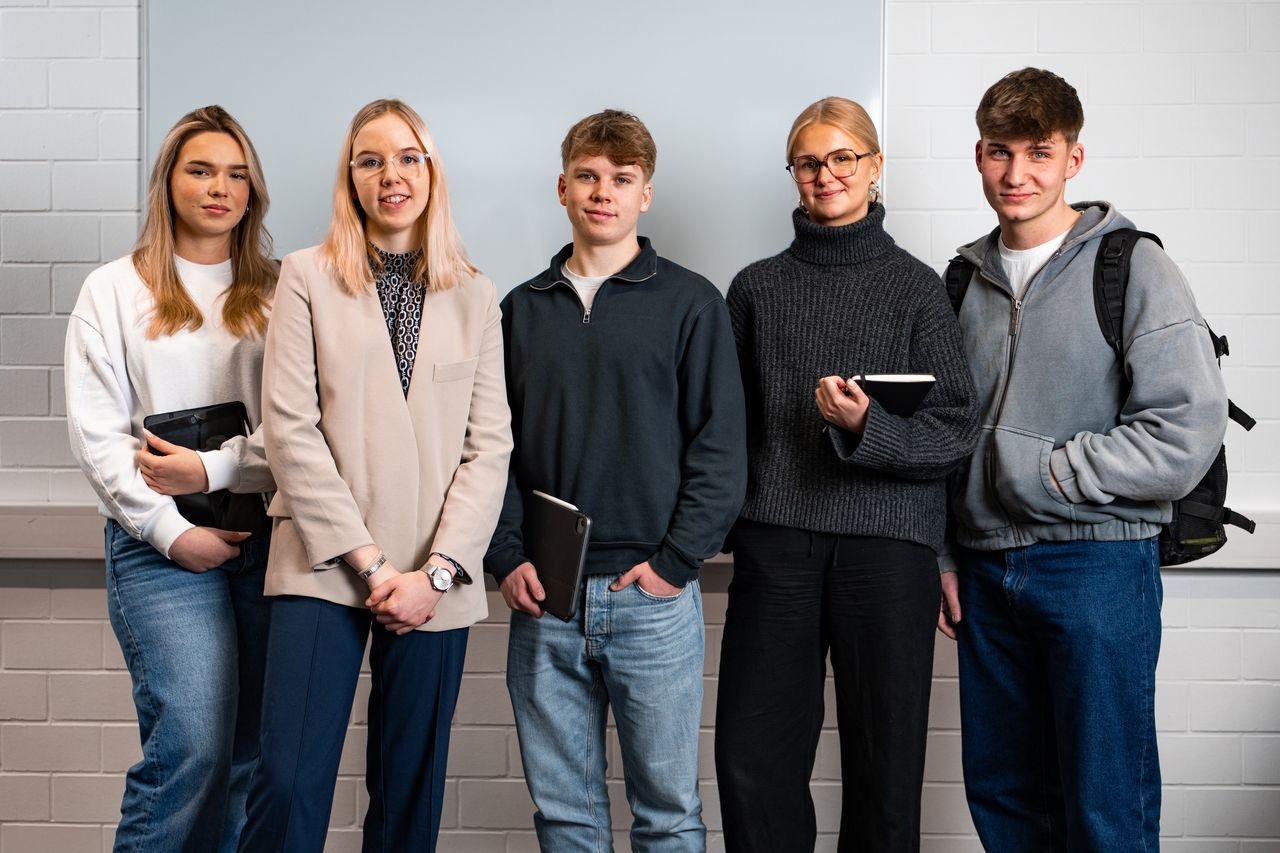 Fünf junge Studierende stehen nebeneinander vor einer Backsteinwand und halten Tablets und Notizbücher in den Händen