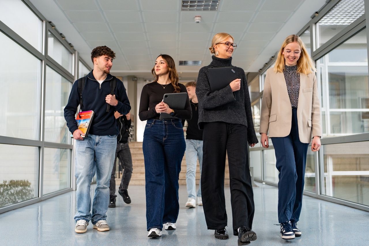Studierende gehen lachend durch einen hellen Hochschulflur, mit Laptop, Tablet und Büchern in der Hand