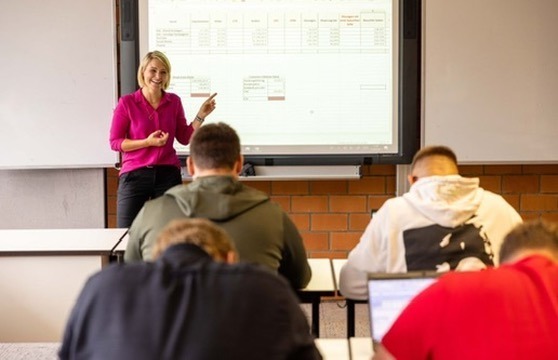 Dozentin zeigt auf eine Tabelle an der Leinwand, während Studierende im Hörsaal zuhören