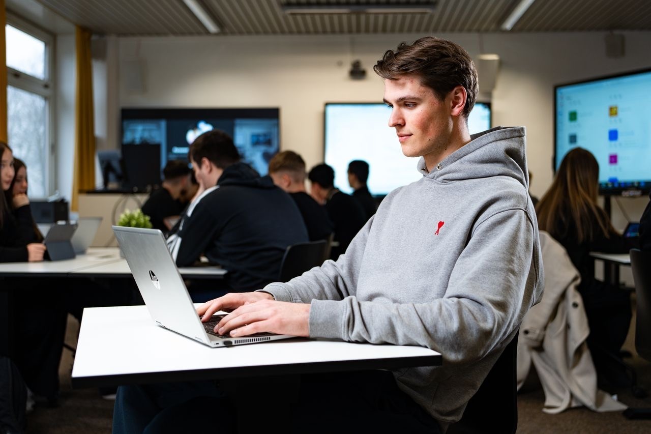 Junger Mann arbeitet an einem Laptop in einem modernen Klassenraum mit weiteren Schülern im Hintergrund