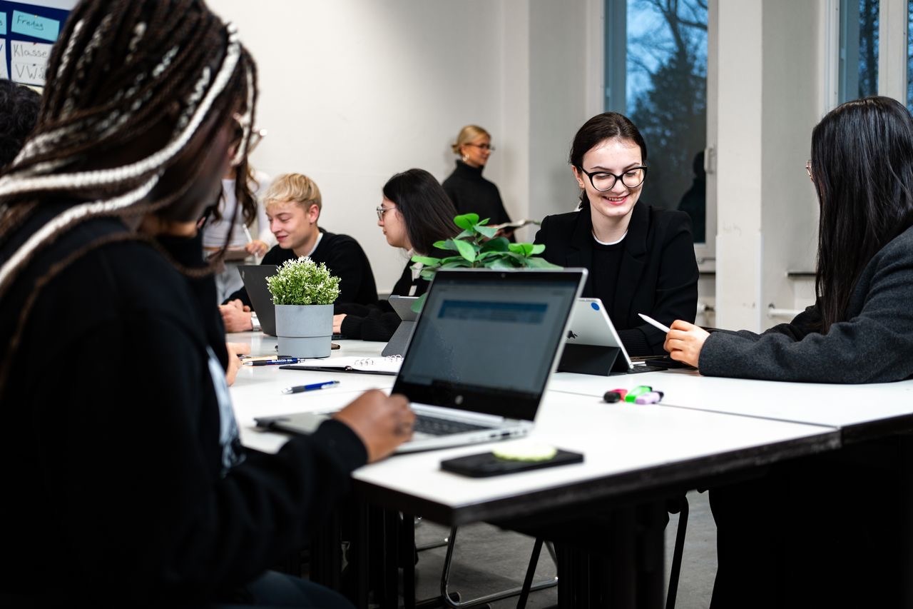 Schülerinnen und Schüler arbeiten gemeinsam an Tischen mit Laptops in einem Klassenzimmer, eine Lehrerin steht im Hintergrund