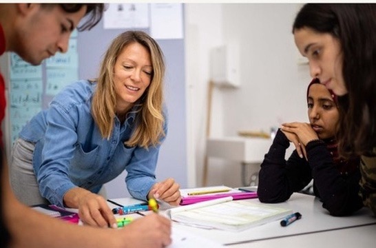 Eine Lehrerin beugt sich lächelnd über einen Tisch und unterstützt drei Schülerinnen beim Lernen im Unterricht