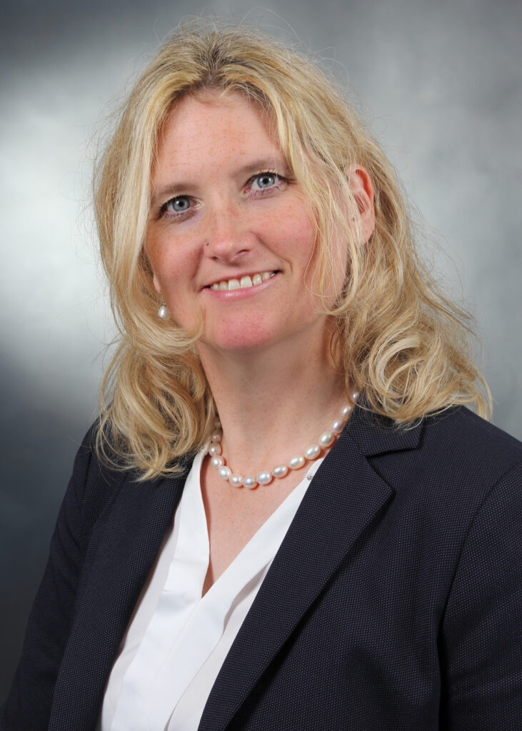 Porträtfoto einer lächelnden Frau mit blonden Haaren, blauem Blazer und Perlenkette