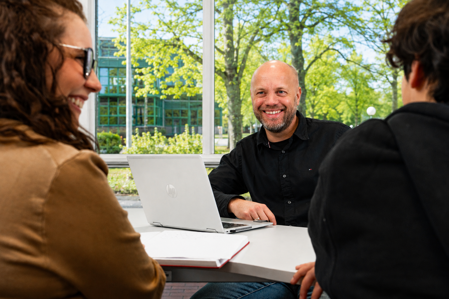 Drei Personen in einem freundlichen Gespräch an einem Tisch mit Laptop, im Hintergrund ein begrünter Campus