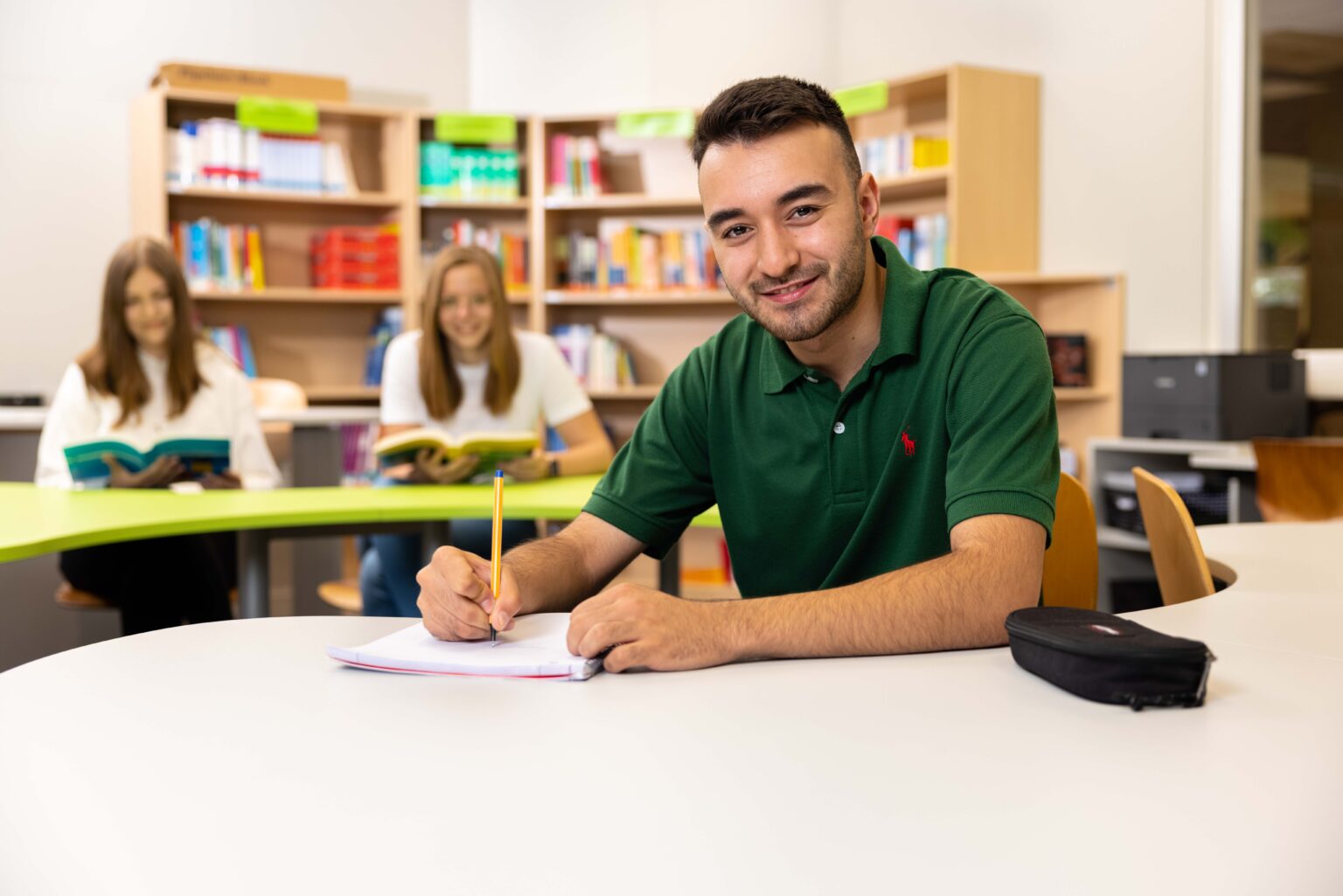Lächelnder junger Mann schreibt in einem Notizbuch in einer Schulbibliothek, im Hintergrund zwei lernende Mitschülerinnen