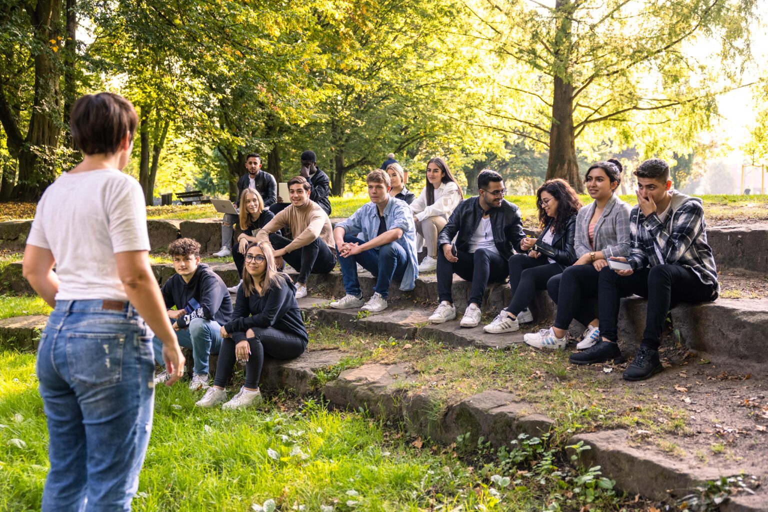 Gruppe junger Menschen sitzt auf Steinstufen im Park und hört einer Person zu, die vor ihnen steht